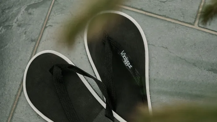 Schwarze Flip-Flops mit weißem Rand auf grauen Steinfliesen, teilweise von Pflanze verdeckt